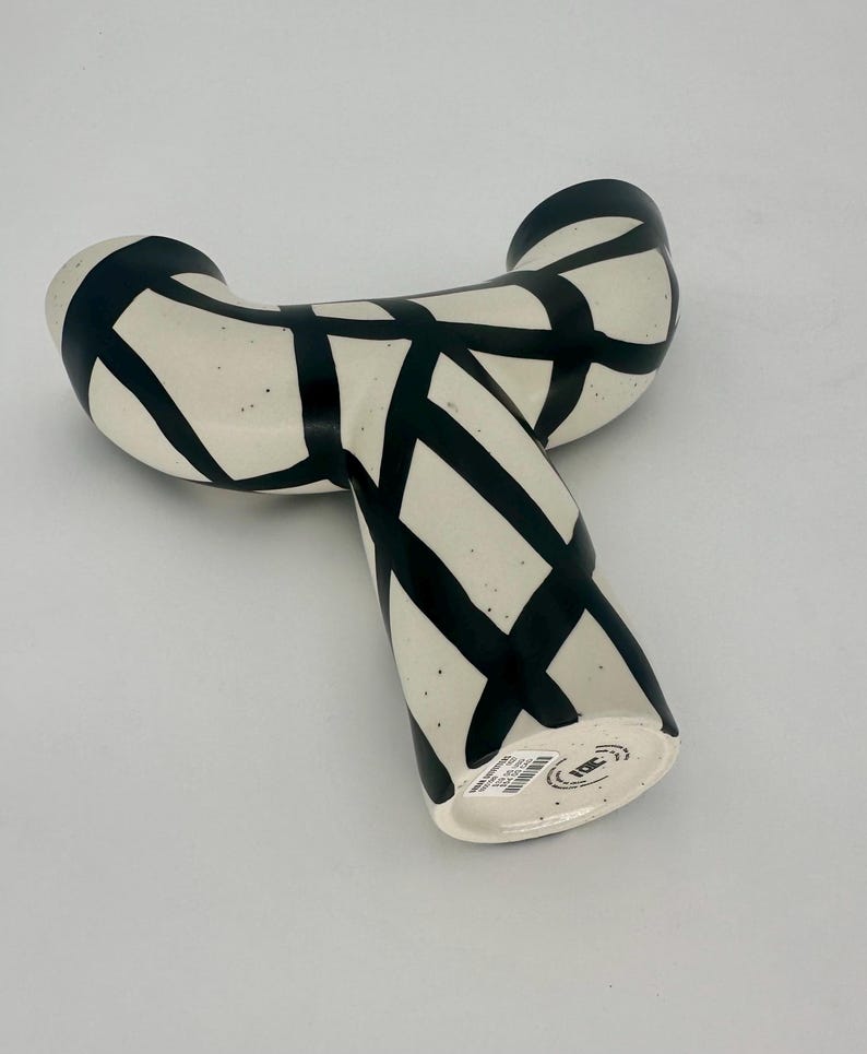 May include: A cream-colored ceramic vase with a unique, curved shape. The vase is decorated with bold black intersecting lines, creating a striking geometric pattern. The vase has a speckled texture and a modern aesthetic.