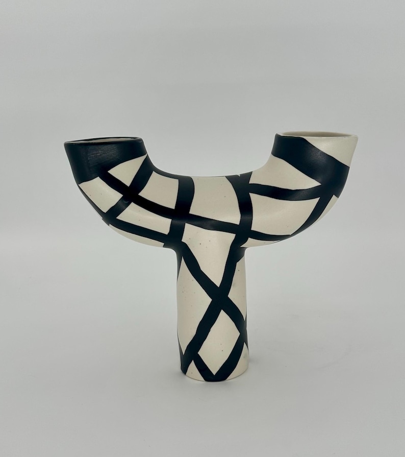 May include: A cream-colored ceramic vase with a unique Y-shape design. The vase features black geometric patterns and accents. The top and the base of the vase are black. The vase is a decorative item.