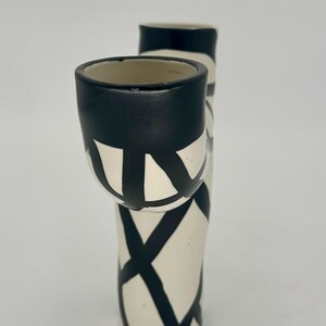 May include: A ceramic vase with a unique design. The vase is primarily off-white with bold black geometric patterns. The top of the vase and the rim of the attached cup are painted black. The vase has a modern, artistic aesthetic.