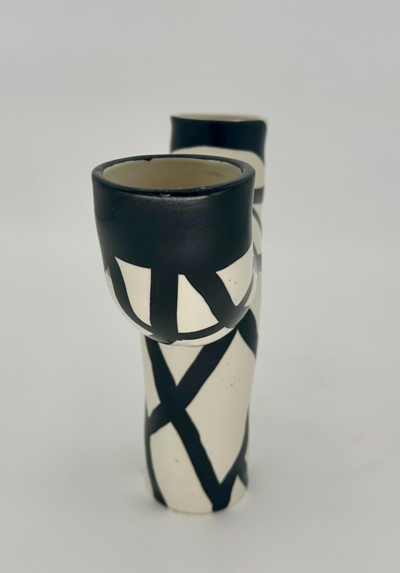 May include: A ceramic vase with a unique design. The vase is primarily off-white with bold black geometric patterns. The top of the vase and the rim of the attached cup are painted black. The vase has a modern, artistic aesthetic.