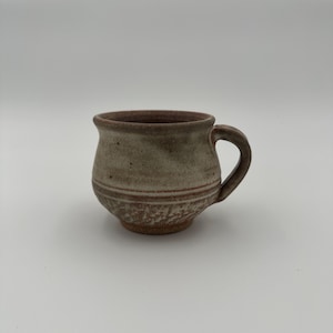 May include: A small, brown and white ceramic mug with a rounded body and a handle. The mug has a rustic, handmade look.