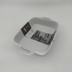 May include: A white rectangular Staub ceramic baking dish with two handles. A product tag is attached to the side. The dish is empty and sits on a white surface.