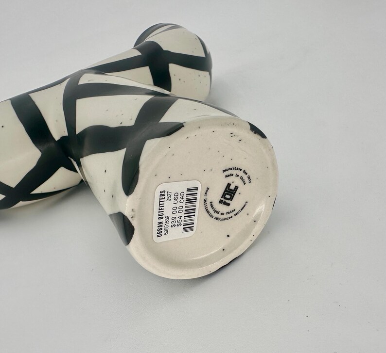 May include: Two ceramic vases with a cream-colored base and black geometric designs. The vases are cylindrical with a slightly flared top. The base of one vase has a sticker from Urban Outfitters.