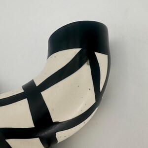 May include: A curved ceramic vase with a black and white geometric pattern. The vase has a black rim and a cream-colored body. The black pattern consists of intersecting lines that create a grid-like design.