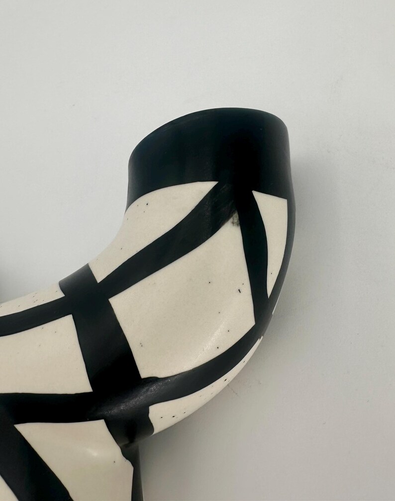 May include: A curved ceramic vase with a black and white geometric pattern. The vase has a black rim and a cream-colored body. The black pattern consists of intersecting lines that create a grid-like design.