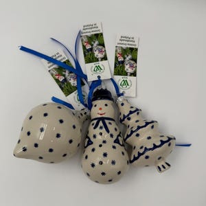 May include: Three ceramic Christmas ornaments with blue star patterns on a cream background. One is a teardrop shape, one is a snowman with a blue scarf, and one is a tiered tree shape. Each has a blue ribbon and a tag reading Christmas Ornament Handmade in Poland.