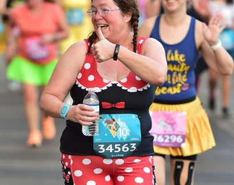 May include: A woman wearing a red and white polka dot outfit with a black top and a Minnie Mouse bow. She is holding a water bottle and has a race bib with the number 34563 on it. She is giving a thumbs up and smiling.
