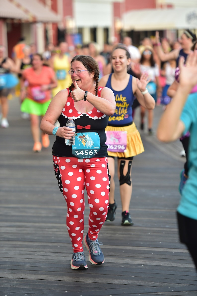 May include: A woman wearing a red and white polka dot outfit with a black top and a Minnie Mouse bow. She is holding a water bottle and has a race bib with the number 34563 on it. She is giving a thumbs up and smiling.