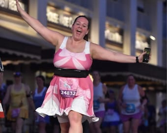 May include: A woman wearing a pink running dress with a large pink bow on the front. She is holding a cell phone in her right hand and has a race bib with the number 24832 on it.