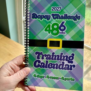 May include: A spiral-bound training calendar with a green and purple plaid design. The cover reads "2027 Dopey Challenge 48.6 miles Training Calendar" with text in green and purple. The calendar is held by a person.