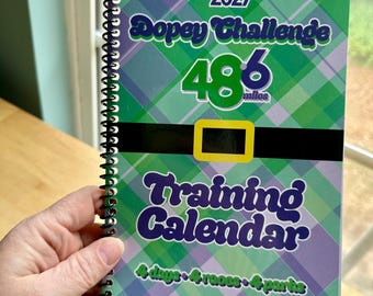 May include: A spiral-bound training calendar with a green and purple plaid design. The cover reads "2027 Dopey Challenge 48.6 miles Training Calendar" with text in green and purple. The calendar is held by a person.