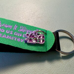 May include: A purple and green charm with the number "2180" on it. The charm is attached to a black and green strap with the text "Dream It, Sleep It, Do Us on E, Team It" in purple.