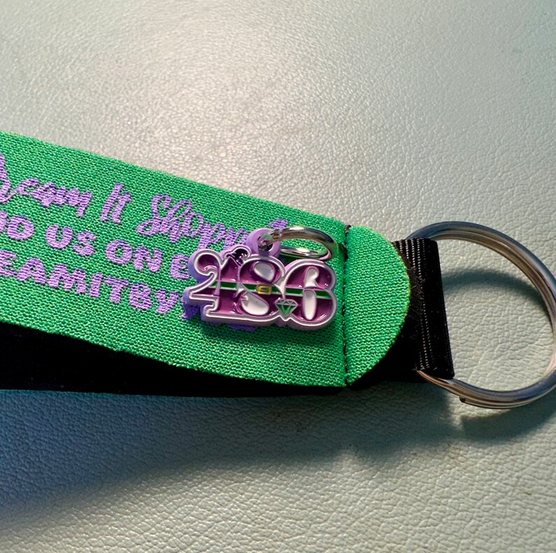 May include: A purple and green charm with the number "2180" on it. The charm is attached to a black and green strap with the text "Dream It, Sleep It, Do Us on E, Team It" in purple.
