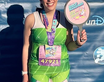 May include: A woman wearing a green and white running outfit with a pink and purple medal around her neck. She is holding a pink sign that says "Half Marathon 13.1 I Did It!" with a cartoon duck on it. The sign is attached to a black handle.