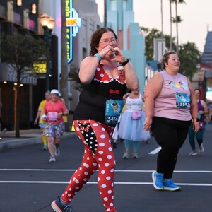 May include: A woman wearing a black and white polka dot shirt and leggings with a red bow and a Minnie Mouse head. She is holding a water bottle and making a heart shape with her hands. She is wearing a race bib with the number 34563. She is running in a race.