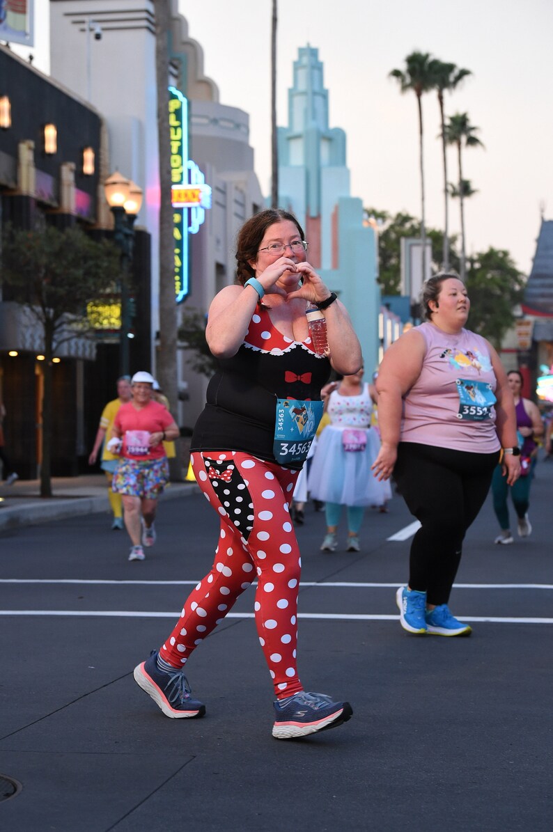 May include: A woman wearing a black and white polka dot shirt and leggings with a red bow and a Minnie Mouse head. She is holding a water bottle and making a heart shape with her hands. She is wearing a race bib with the number 34563. She is running in a race.