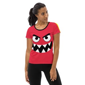 May include: A red athletic t-shirt featuring a cartoon monster face with white eyes, black eyebrows, and a jagged white and black mouth. The shirt has a black neckline and a yellow stripe on the shoulder.
