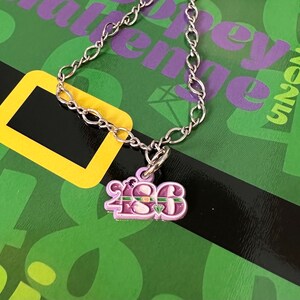 May include: A silver chain necklace with a purple charm that says "1986" with a green and gold stripe across the top. The charm has a small green gem on it.