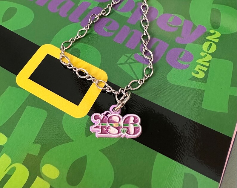 May include: A silver chain necklace with a purple charm that says "1986" with a green and gold stripe across the top. The charm has a small green gem on it.