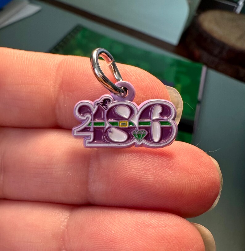 May include: A purple metal charm with the number "2183" in white lettering. The charm has a green and white band around the numbers and a small diamond in the center.