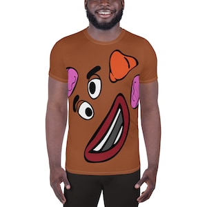 Puede incluir: Una camiseta marrón con una cara de dibujos animados impresa en la parte delantera. La cara tiene dos ojos grandes, una amplia sonrisa y dos orejas rosas. La camiseta es de ajuste deportivo y tiene mangas cortas.