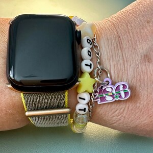 May include: A black Apple Watch with a brown and beige woven band. The watch is worn on a person's wrist with a charm bracelet featuring white and yellow beads, a star charm, and a purple charm with the text "2023".
