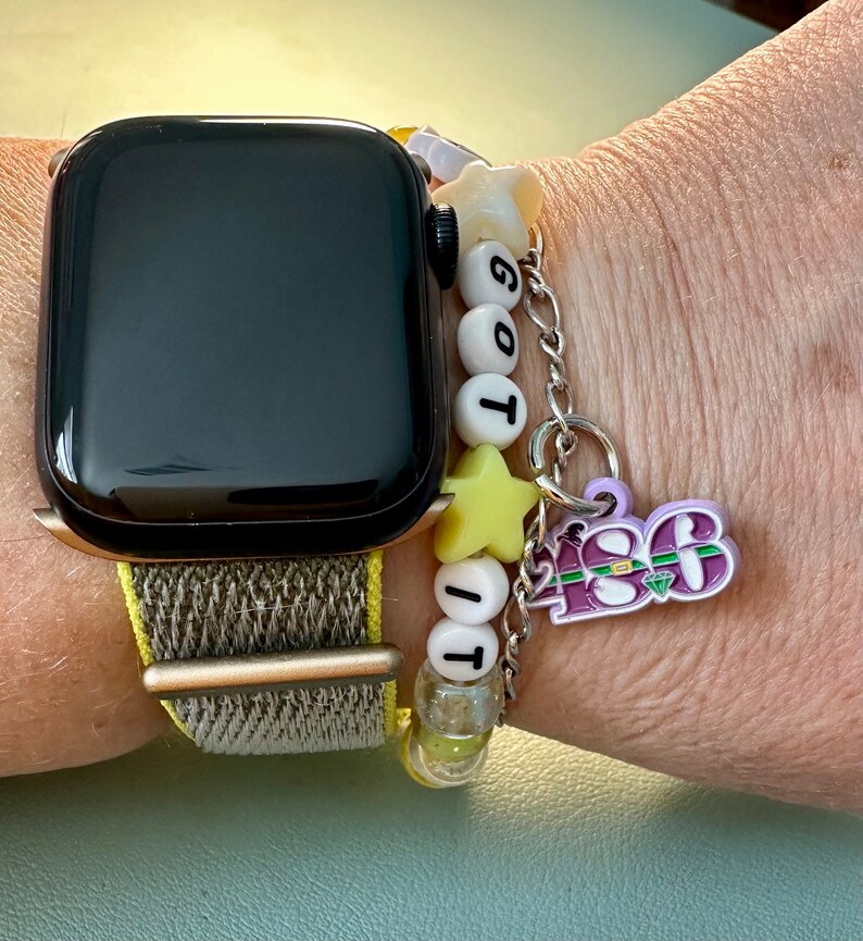 May include: A black Apple Watch with a brown and beige woven band. The watch is worn on a person's wrist with a charm bracelet featuring white and yellow beads, a star charm, and a purple charm with the text "2023".