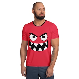 May include: Red t-shirt with a black and white monster face graphic. The graphic features white eyes, black eyebrows, and a jagged black mouth. The shirt has a black trim around the collar.