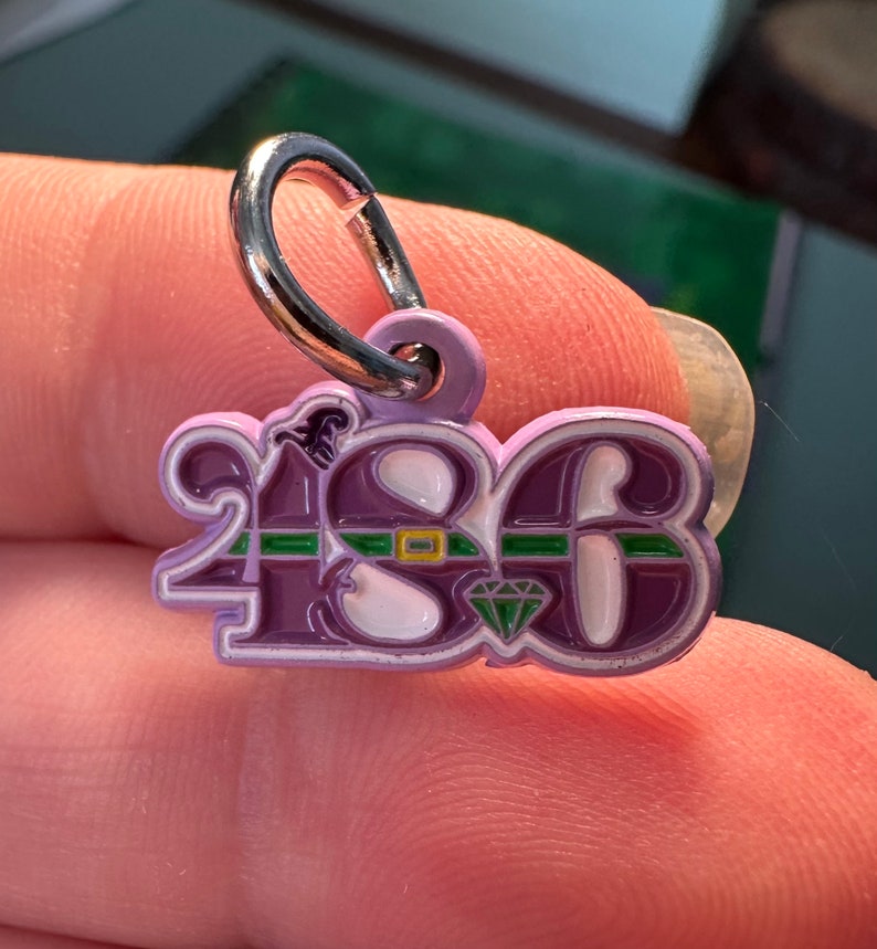 May include: Purple metal charm with the number 2183, a green and yellow belt, and a diamond. The charm has a silver loop for attaching to a necklace or bracelet.