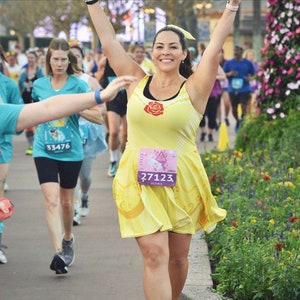 May include: A woman wearing a yellow running dress with a red rose and a pink race bib with the number 27123. She is running in a race with other people.