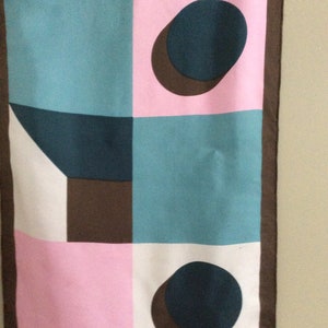 May include: A rectangular scarf with a geometric pattern. The design features squares and circles in teal, pink, white, and brown. The scarf has a dark brown border.