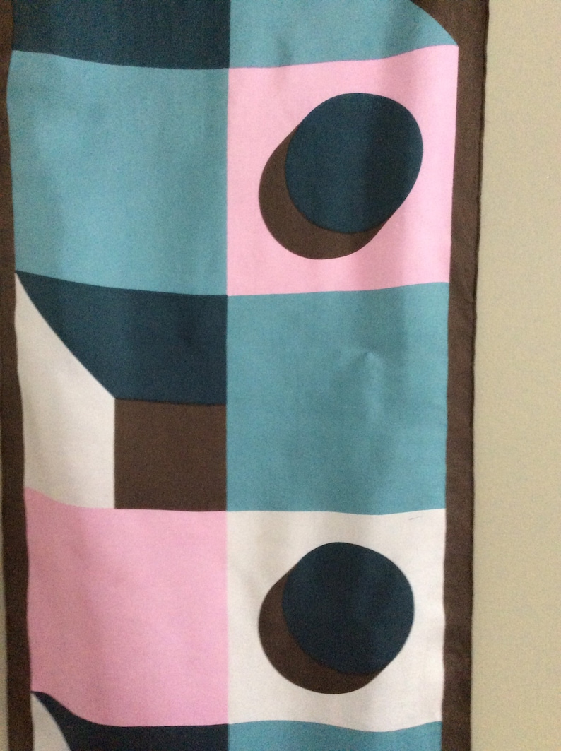 May include: A rectangular scarf with a geometric pattern. The design features squares and circles in teal, pink, white, and brown. The scarf has a dark brown border.