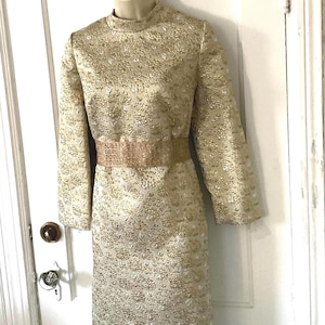 May include: A gold and white brocade dress with a high neckline and long sleeves. The dress has a matching belt.