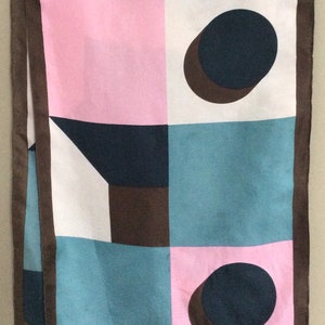 May include: A rectangular scarf with a geometric pattern in pink, white, teal, and brown. The scarf features a repeating design of squares, circles, and geometric shapes. The scarf is trimmed with a brown border and has the text "Jacques Fath" in the corner.