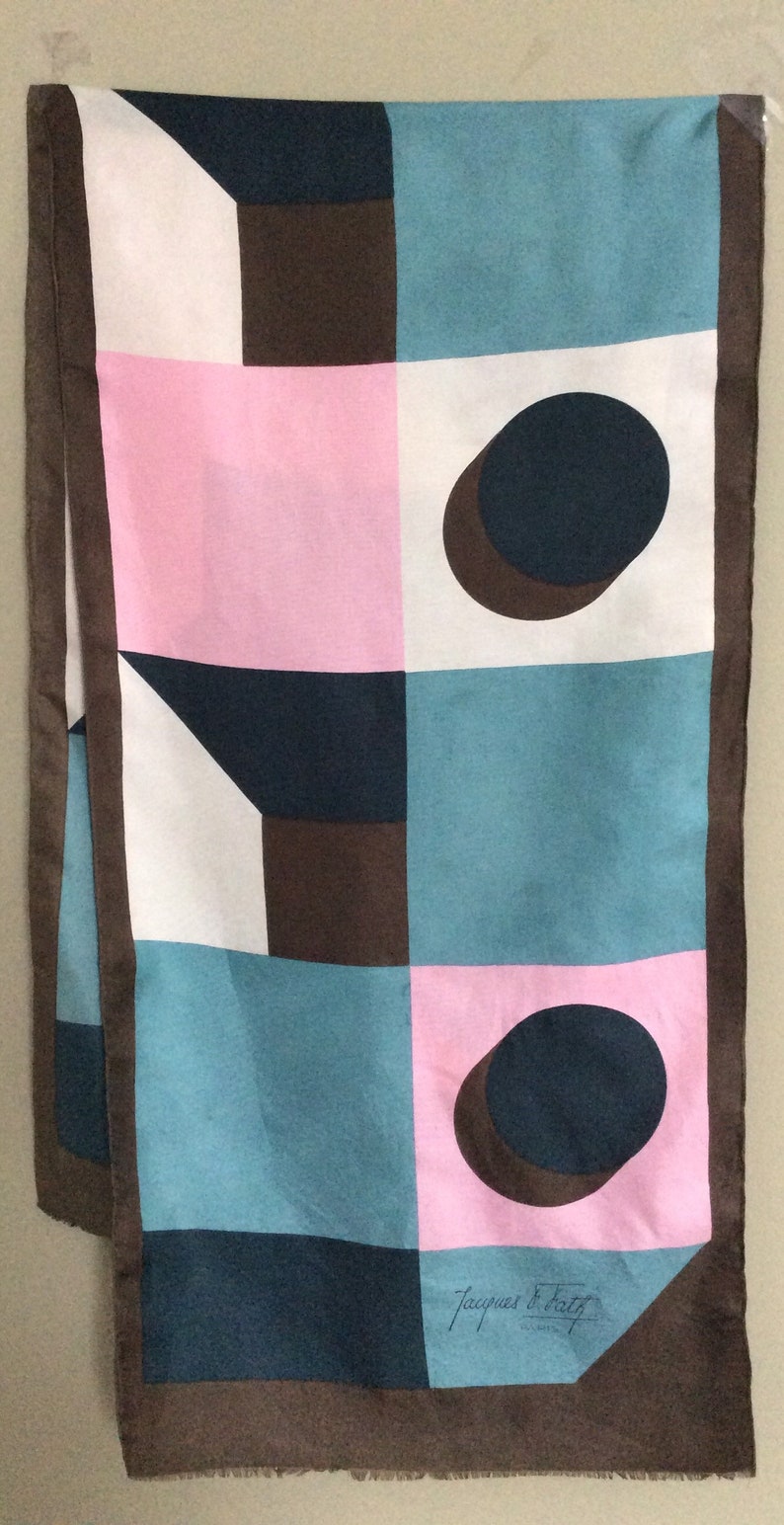May include: A rectangular scarf with a geometric pattern in pink, white, teal, and brown. The scarf features a repeating design of squares, circles, and geometric shapes. The scarf is trimmed with a brown border and has the text "Jacques Fath" in the corner.
