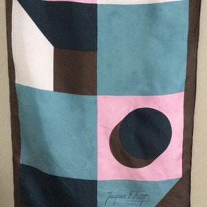 May include: A vintage scarf with a geometric design in pink, teal, white, and brown. The scarf features a repeating pattern of squares, circles, and triangles. The Jacques Fath Paris signature is visible.