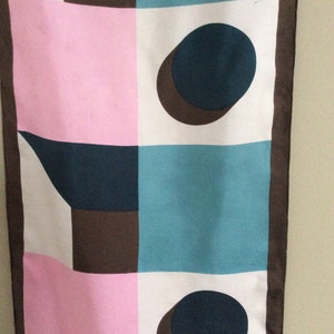 May include: A rectangular scarf with a geometric pattern. The design features squares and shapes in pink, teal, white, brown, and navy blue. Two navy blue circles with brown shadows are present. The scarf has a brown border.