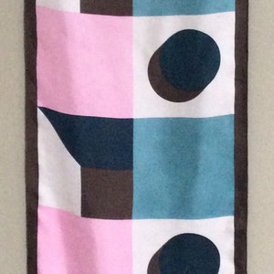 May include: A rectangular scarf with a geometric pattern. The design features squares and shapes in teal, pink, white, and brown, with dark brown ovals. The scarf is bordered with a dark brown trim.
