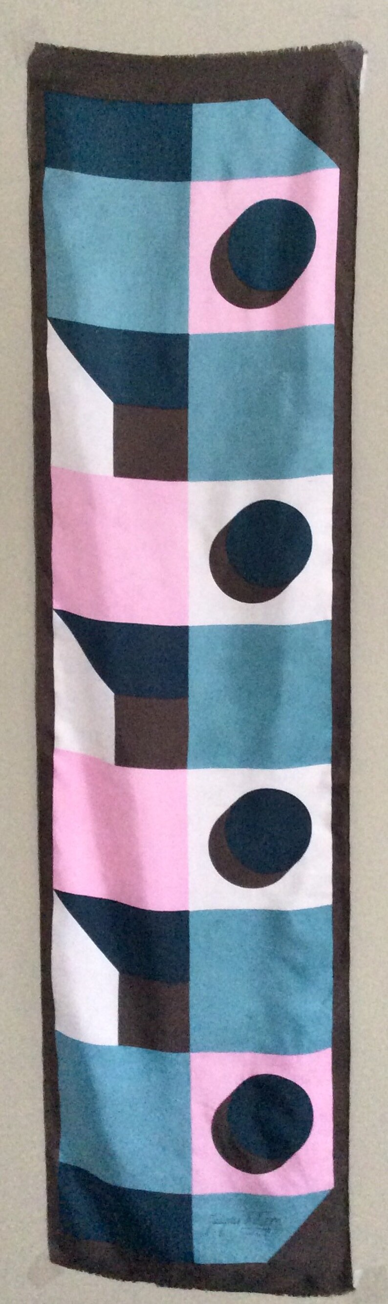 May include: A rectangular scarf with a geometric pattern. The design features squares and shapes in teal, pink, white, and brown, with dark brown ovals. The scarf is bordered with a dark brown trim.