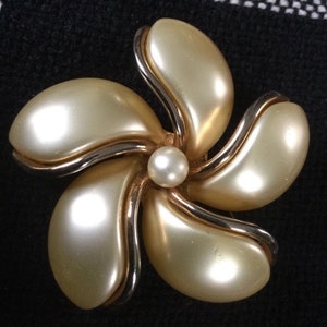 May include: A vintage flower-shaped brooch with five creamy-colored petals and a pearl center. The petals are outlined with a silver-colored metal and have a gold-colored metal border. The brooch is set against a dark background.