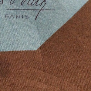 May include: A section of a silk scarf with a light blue and brown color scheme. The scarf has the text "Jacques O' Valz PARIS" in black script. The scarf is folded, showing a geometric pattern.