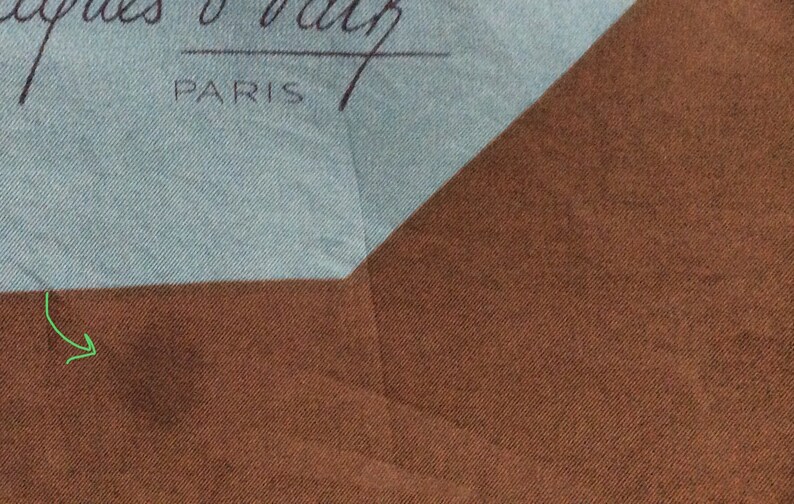 May include: A section of a silk scarf with a light blue and brown color scheme. The scarf has the text "Jacques O' Valz PARIS" in black script. The scarf is folded, showing a geometric pattern.