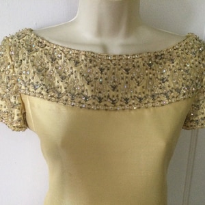 May include: A pale yellow dress with a beaded neckline and cap sleeves. The neckline is heavily embellished with silver and clear beads, creating a sparkling effect. The dress has a simple, elegant design.