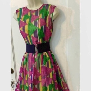 May include: A sleeveless, patterned dress with a vibrant mix of colors, including pink, green, yellow, and purple. The dress features a geometric design and is cinched at the waist with a wide black belt. The dress is displayed on a mannequin.