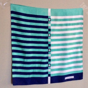 May include: A turquoise and white striped silk scarf with a navy blue border. The scarf has a geometric pattern of white squares and diamonds on the right side. The scarf is labeled "Givenchy" in the bottom right corner.