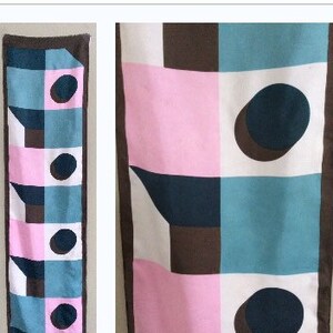 May include: A rectangular scarf with a geometric pattern. The design features squares of pink, teal, white, and brown, with dark blue circles and angular shapes. The scarf has a brown border.