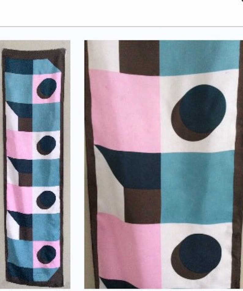 May include: A rectangular scarf with a geometric pattern. The design features squares of pink, teal, white, and brown, with dark blue circles and angular shapes. The scarf has a brown border.