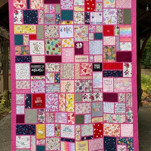 May include: A patchwork quilt featuring a bright pink border and numerous rectangular fabric blocks with diverse patterns, including floral, polka dot, and animal prints. Several blocks contain text such as "DADDY'S mini," "BEST FRIEND," and "PARTNER IN CRIME."