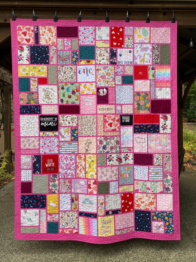 May include: A patchwork quilt featuring a bright pink border and numerous rectangular fabric blocks with diverse patterns, including floral, polka dot, and animal prints. Several blocks contain text such as "DADDY'S mini," "BEST FRIEND," and "PARTNER IN CRIME."