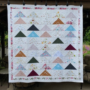 May include: A rectangular quilt featuring a repeating pattern of solid and floral-printed triangles arranged in a grid. The quilt has a white background with a floral border and is constructed with visible stitching throughout the fabric surface.
